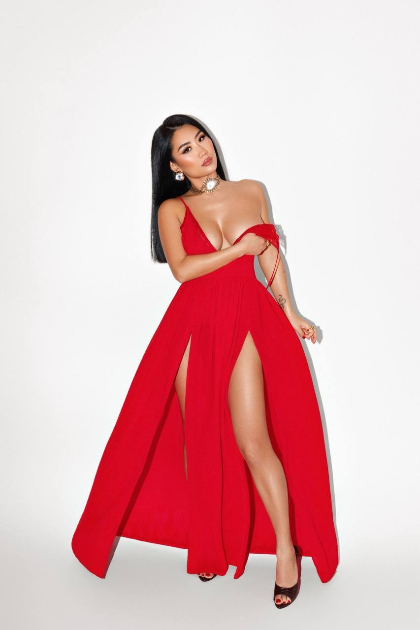 Elegant brunette luxury escort model wearing a flowing red evening gown with dramatic high slits, minimalist white studio, haute couture editorial photography