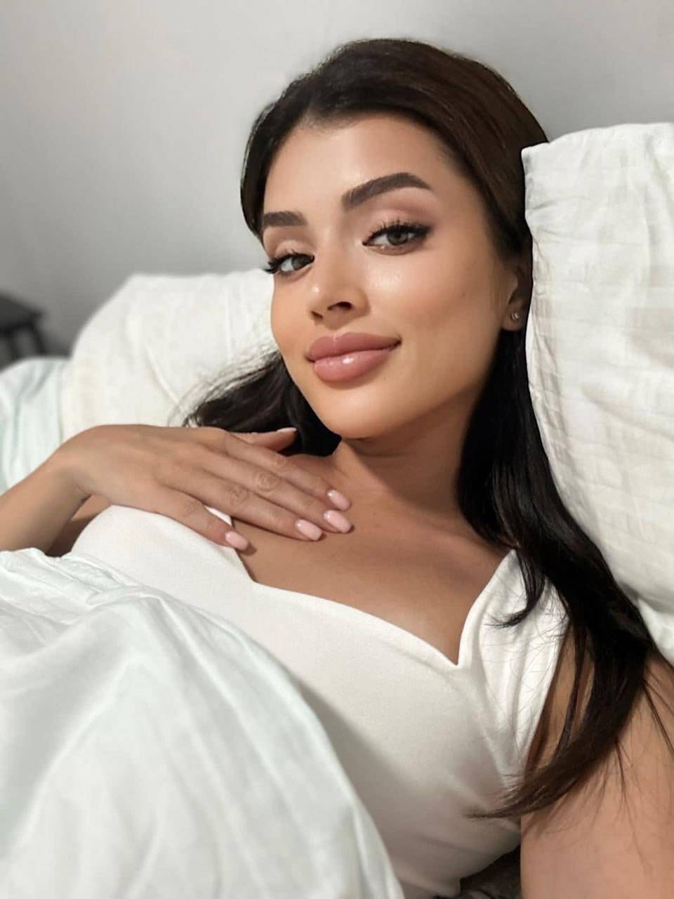 close up portrait of beautiful brunette woman relaxing in bed wearing white satin top, minimal bedroom, soft daylight, natural beauty, cozy aesthetic