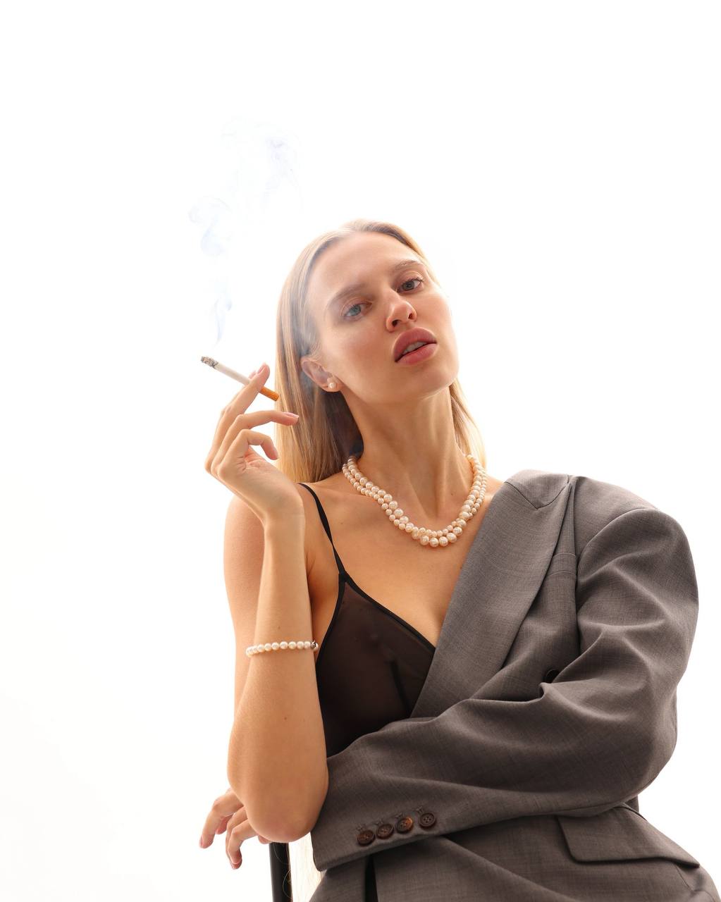 Elegant blonde model wearing a gray tailored blazer over sheer black lingerie with pearl necklace and bracelet, minimalist luxury styling, white seamless background, soft editorial lighting, sophisticated fashion magazine cover, ultra-realistic, premium photography