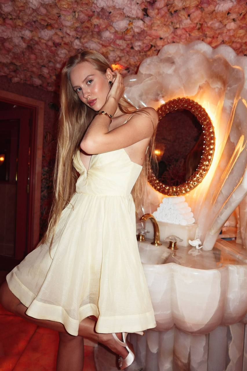 Blonde fashion model with long blonde hair wearing an ivory mini dress posing in a luxury floral bathroom with shell mirror
