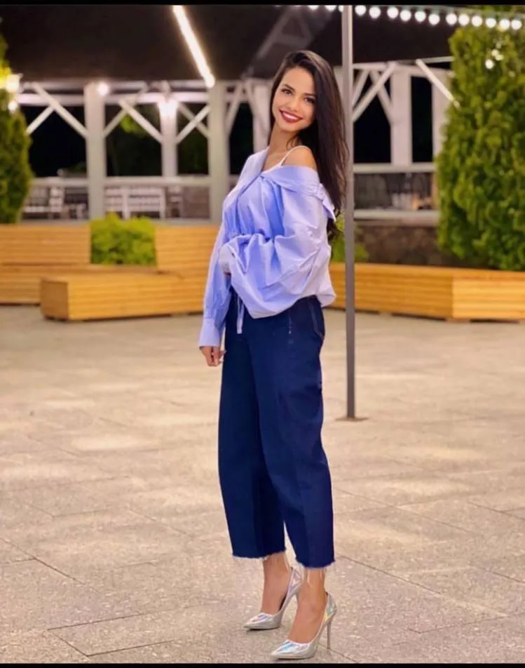smiling brunette woman in oversized light blue off shoulder blouse, navy cropped trousers and silver high heels, modern outdoor plaza at night, city lights, lifestyle fashion photography