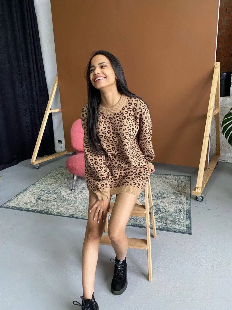 happy brunette woman wearing oversized leopard print sweater and black combat boots, cozy photo studio with warm brown backdrop, casual fashion editorial, natural candid smile