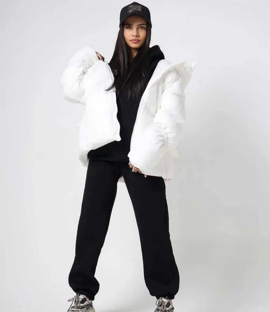 fashion model with long straight black hair wearing oversized white puffer jacket, black hoodie, black joggers and baseball cap, minimal white studio background, modern streetwear editorial photography