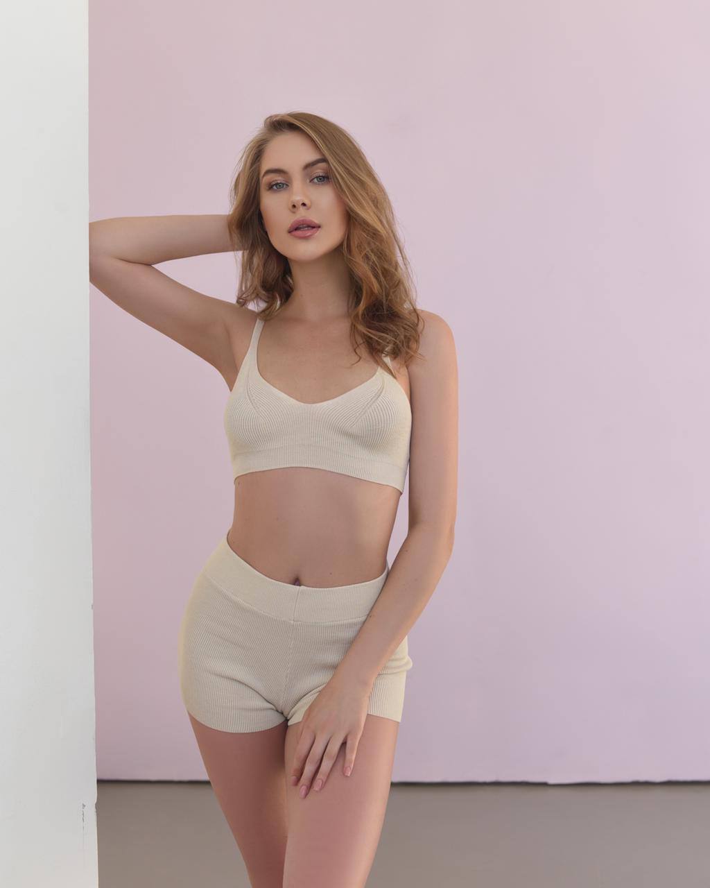 Blonde woman in a cream ribbed knit bralette and shorts set leaning against a white wall on a soft pink background