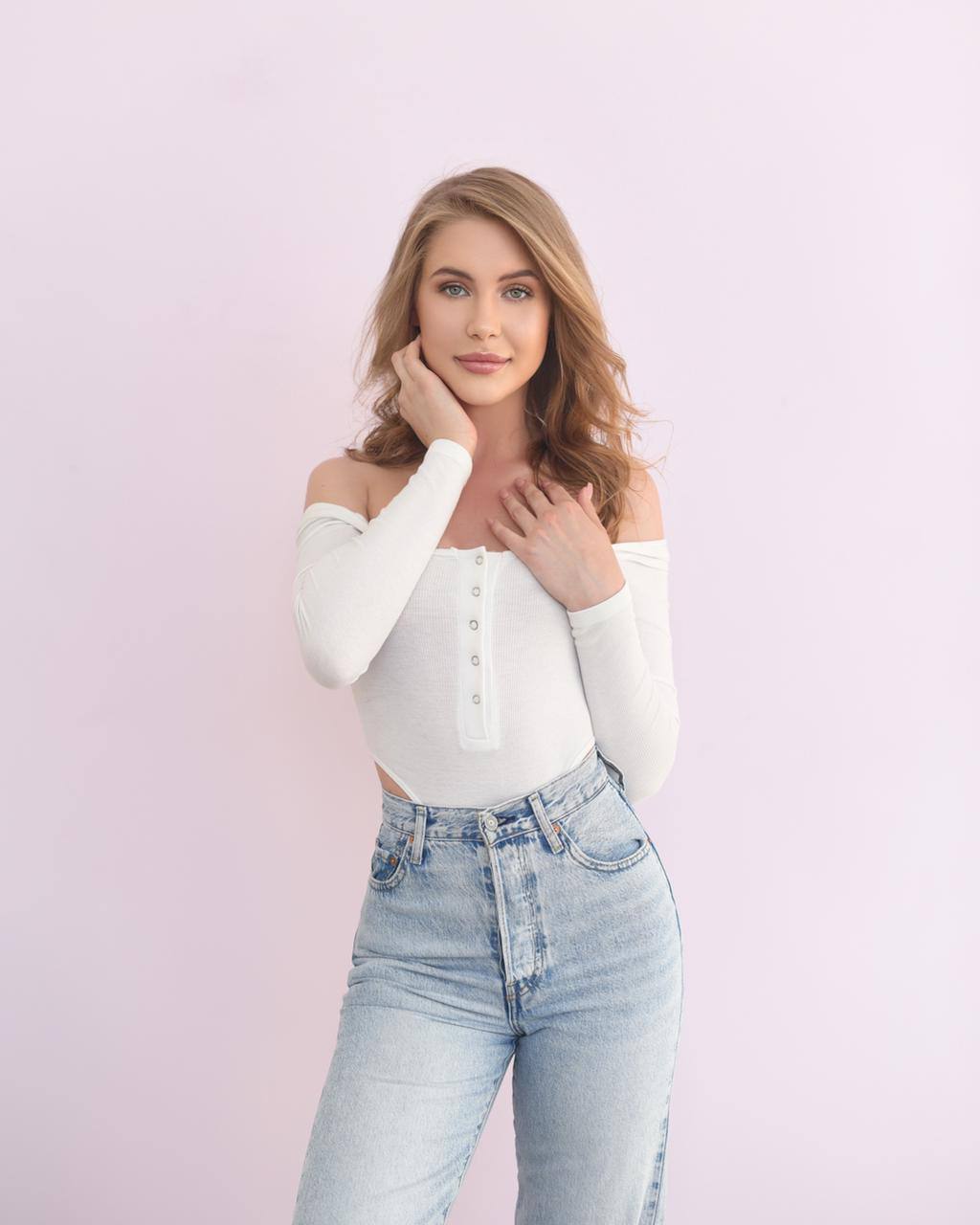 Young woman with wavy blonde hair in a white off-shoulder top and high-waisted light blue jeans on a pale pink background