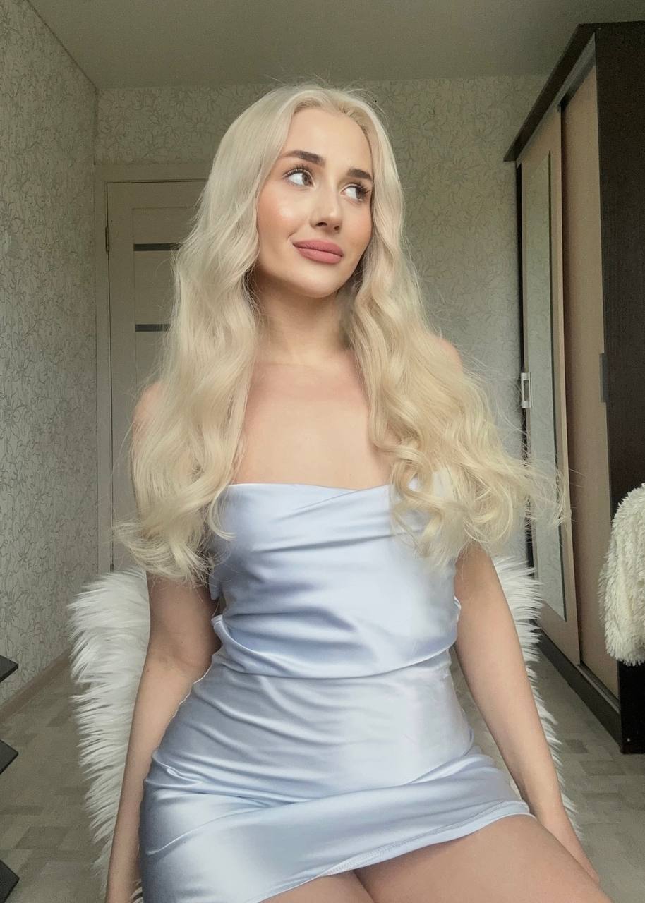 elegant platinum blonde woman in light blue satin mini dress sitting on white fluffy chair