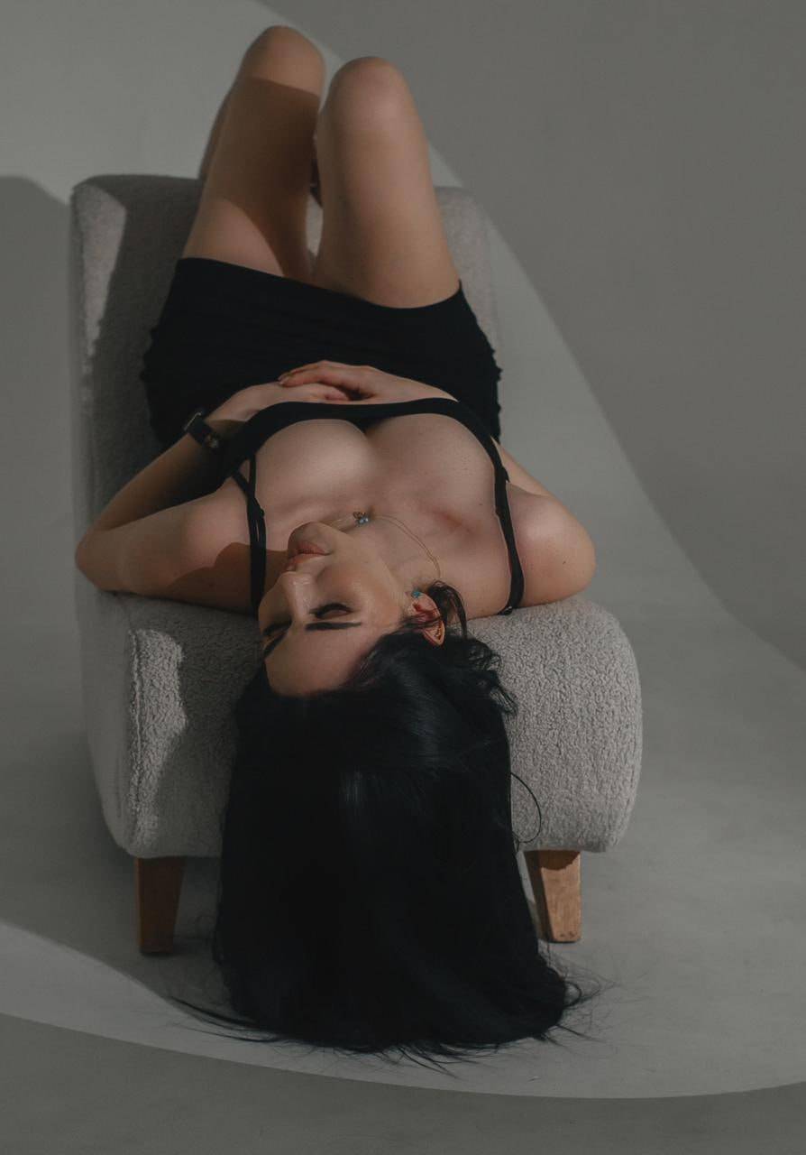 fashion model with long black hair lying on modern armchair wearing little black dress in studio