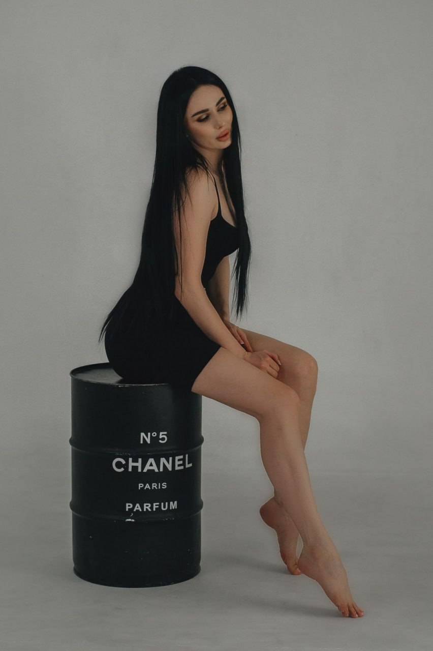 beautiful brunette woman sitting on black Chanel No 5 barrel wearing elegant black mini dress minimalist studio