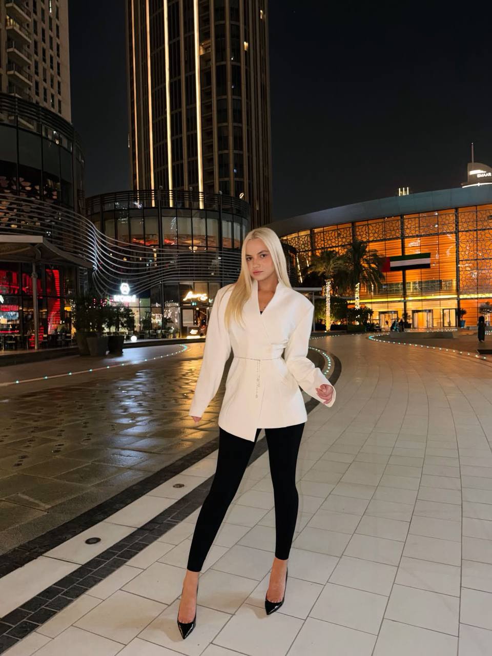 Beautiful blonde fashion model wearing a tailored white blazer, black leggings and black high heels, posing in front of modern Dubai skyscrapers at night, luxury city lights, premium street style editorial photography.