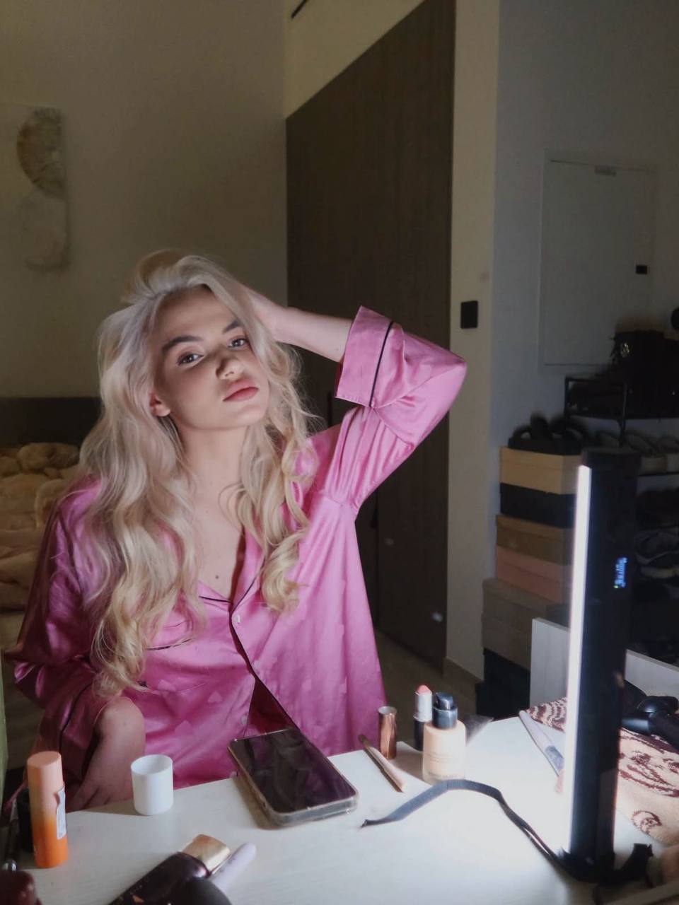 Luxury beauty portrait of a blonde model wearing pink satin silk pajamas, sitting at a vanity table with makeup products, soft indoor lighting, glamorous morning routine, editorial fashion photography.