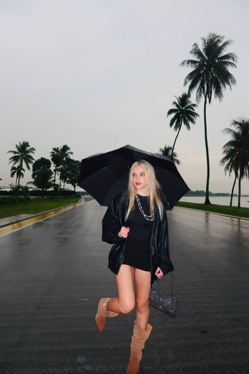 Beautiful blonde fashion model walking under a black umbrella on a rainy palm-lined boulevard, wearing a black mini dress, oversized leather jacket, beige suede boots and carrying a luxury quilted handbag, cinematic rainy atmosphere, editorial street fashion.