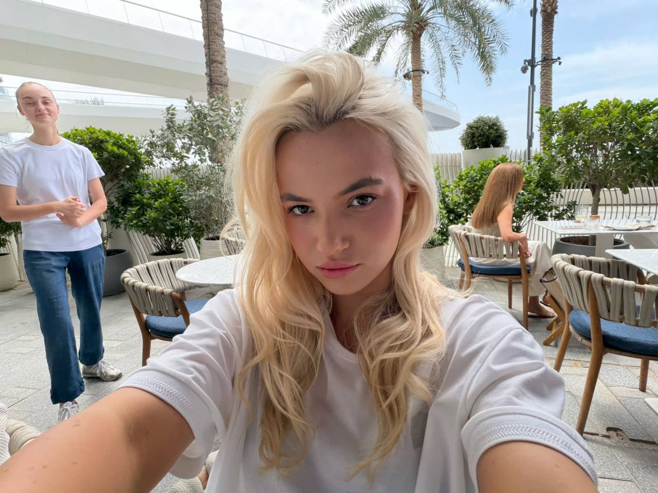 Natural lifestyle selfie of a beautiful blonde model wearing a casual oversized white T-shirt at a luxury outdoor café surrounded by palm trees, soft daylight, minimal makeup, premium social media lifestyle aesthetic.