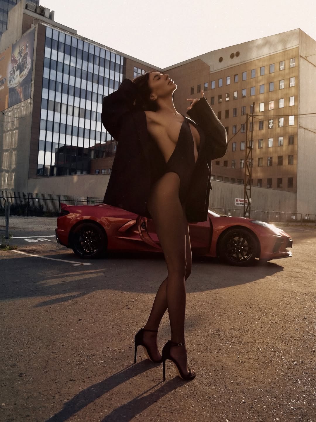 Ultra-realistic luxury fashion model with long silky dark brown hair flowing naturally, flawless bronzed skin, elegant feminine figure, standing confidently in front of a red exotic supercar with the driver's door open, wearing an ultra-deep plunging black bodysuit beneath an oversized black tailored blazer draped loosely over her shoulders, sheer black tights and elegant black stiletto heels, photographed during golden hour in an upscale modern financial district, warm sunset light creating dramatic rim lighting and long cinematic shadows, modern glass skyscrapers surrounding the scene, premium luxury lifestyle aesthetic, confident editorial pose with head tilted back and eyes closed, cinematic composition, luxury fashion campaign, rich golden tones, realistic skin texture, HDR, Sony A7R V, 50mm lens, ultra photorealistic, masterpiece, Vogue editorial, 8k