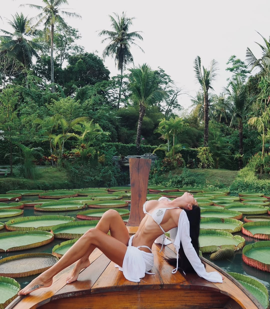 Ultra-realistic luxury travel editorial photography of a breathtaking brunette woman with long silky black hair posing gracefully on the bow of a polished wooden boat surrounded by giant Victoria Amazonica lily pads in a lush tropical botanical garden, wearing an elegant white crystal-embellished bikini with a flowing sheer white cover-up, head tilted back enjoying the warm tropical breeze, toned feminine body, cinematic composition, vibrant emerald jungle, exotic paradise atmosphere, premium resort aesthetic, luxury vacation lifestyle, soft natural daylight, rich tropical colors, Vogue Resort editorial, Leica SL3, 85mm lens, ultra photorealistic, HDR, masterpiece, 8k, realistic skin texture, luxury travel campaign