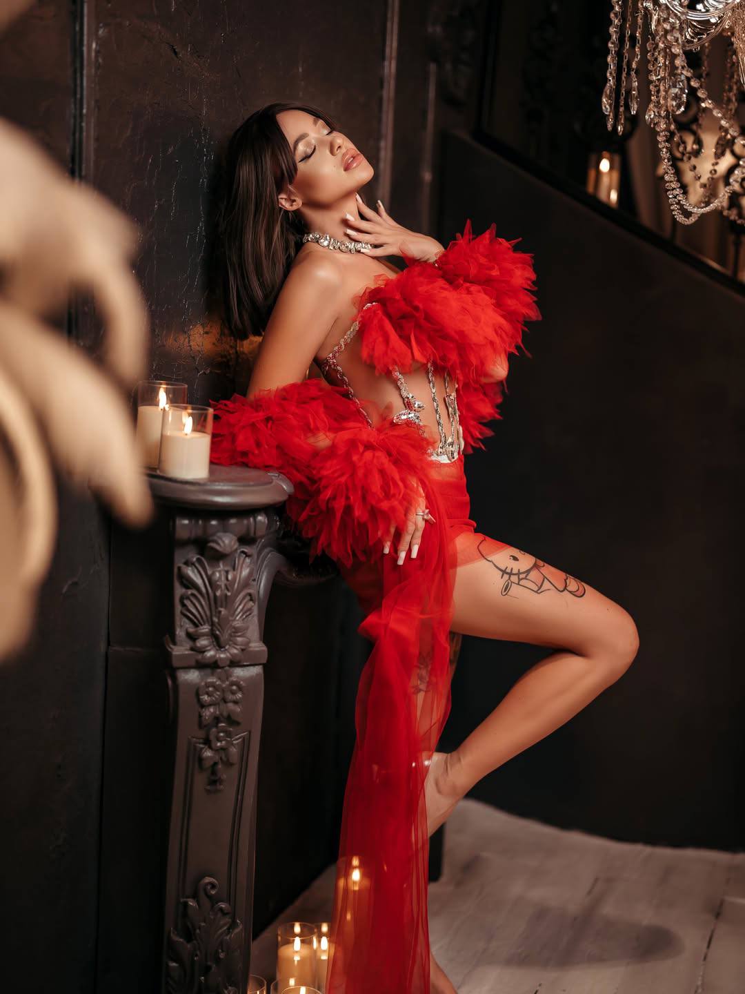 Ultra-realistic luxury editorial fashion photography of an elegant brunette woman with a sleek shoulder-length bob haircut, flawless glowing skin and soft glamorous makeup, leaning gracefully against a dark vintage wall inside an opulent luxury interior, eyes gently closed, wearing an extravagant scarlet couture ensemble with dramatic feathered sleeves, sparkling crystal corset detailing and flowing sheer red tulle train, luxurious diamond choker necklace, long manicured nails, sophisticated sensual pose, ornate black carved pedestal with glowing candles, crystal chandelier details, cinematic warm candlelight mixed with soft studio lighting, rich black and crimson color palette, haute couture, high-fashion Vogue editorial, Dolce & Gabbana Alta Moda aesthetic, luxurious atmosphere, Leica SL3, 85mm Summilux portrait lens, shallow depth of field, ultra photorealistic, HDR, masterpiece, realistic skin texture, premium luxury fashion campaign, 8k