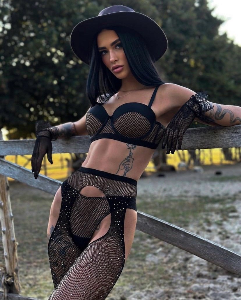 Stylish brunette woman wearing a black mesh lingerie set, wide brim hat and gloves posing outdoors by a wooden fence, bold fashion portrait with luxury styling