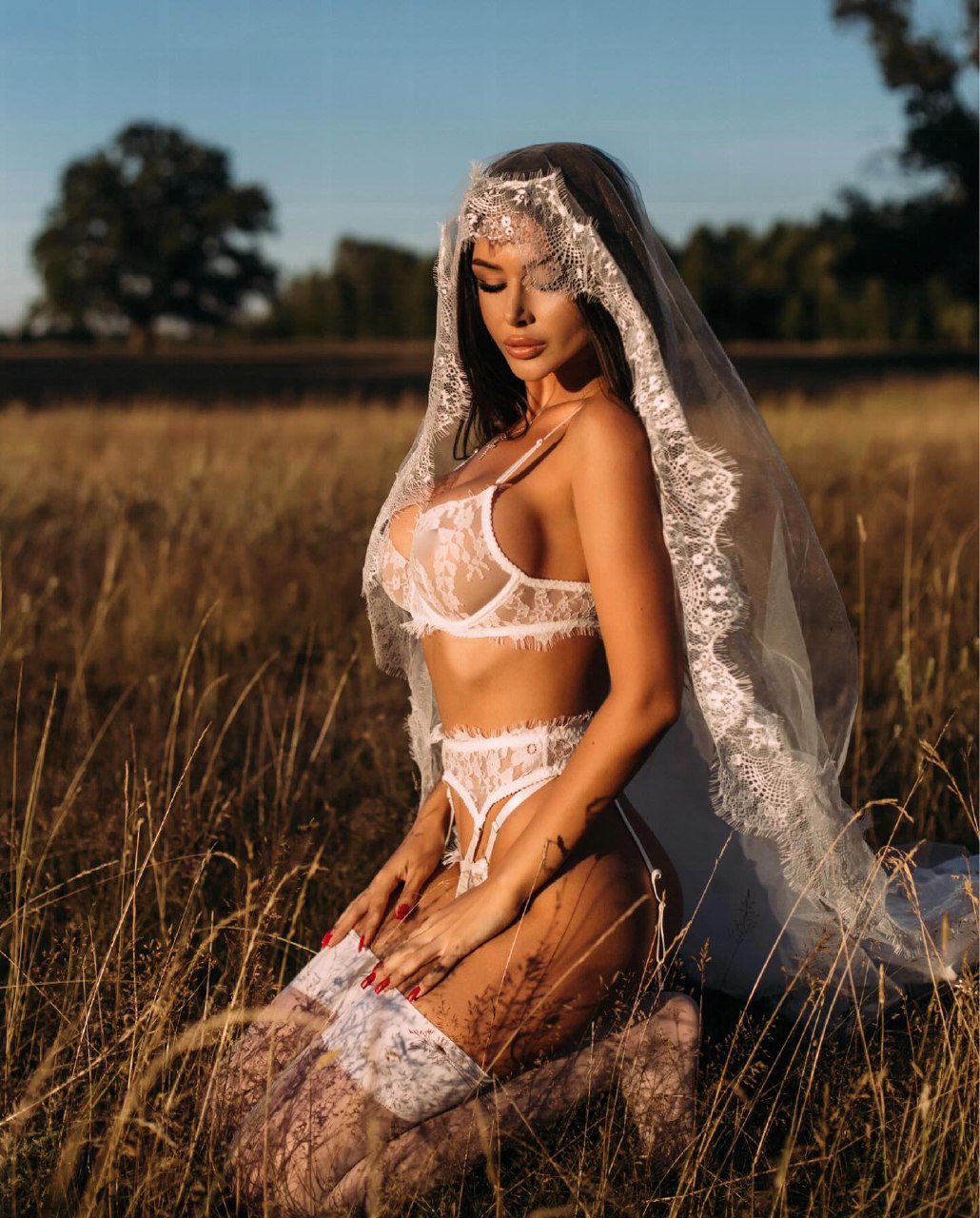Brunette woman wearing elegant white lace bridal lingerie and veil kneeling in a golden field at sunset, luxury bridal boudoir photography with romantic aesthetic