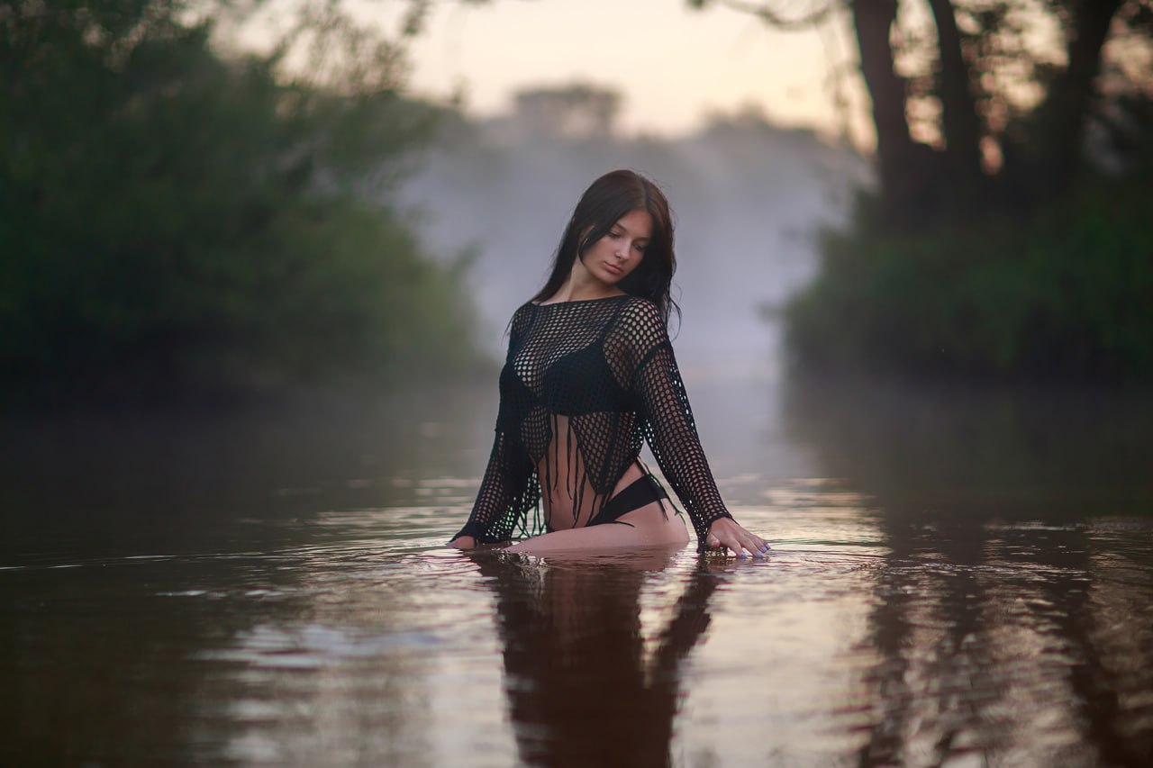 Brunette woman in elegant black mesh beachwear standing in calm river water at sunset, artistic outdoor glamour portrait with natural beauty and serene atmosphere
