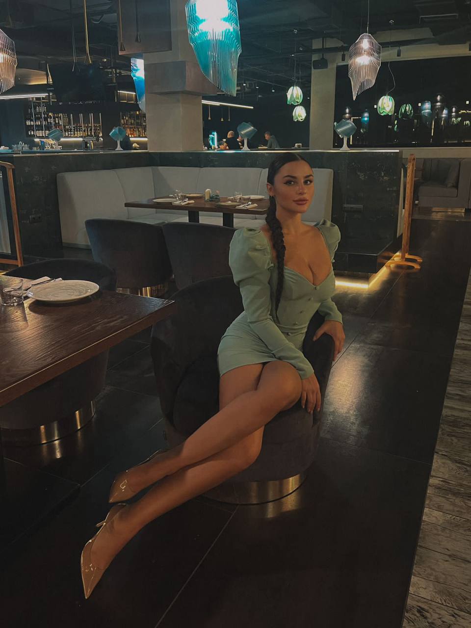 Brunette woman with a long braid in a mint green puff-sleeve mini dress sitting in an armchair at an upscale restaurant in the evening