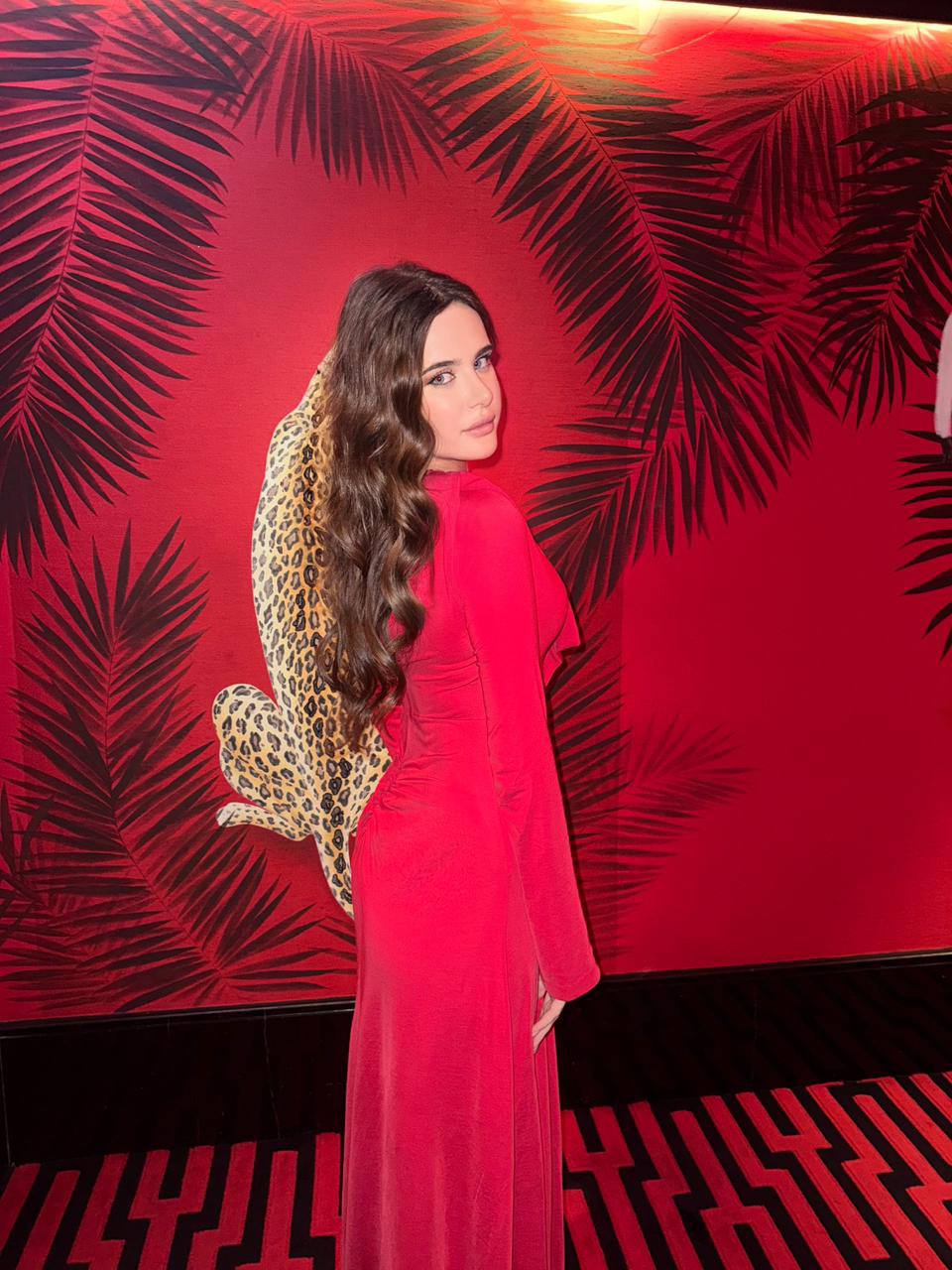 Ultra realistic portrait of a glamorous young brunette woman with long wavy dark brown hair and piercing blue eyes wearing a luxurious red evening gown inside an upscale cocktail lounge with tropical decor, warm ambient lighting, luxury nightlife photography, photorealistic.