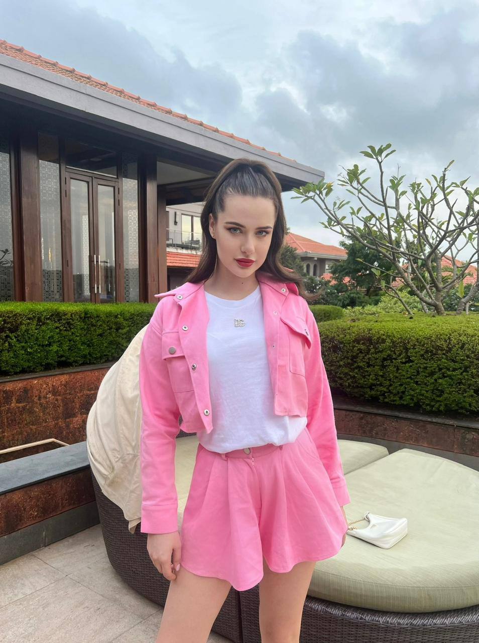 Ultra realistic full body portrait of a fashionable young brunette woman with long dark brown hair tied in a high ponytail and bright blue eyes wearing a stylish pink cropped jacket with matching skirt at a luxury tropical resort villa, premium travel lifestyle photography, 8k.