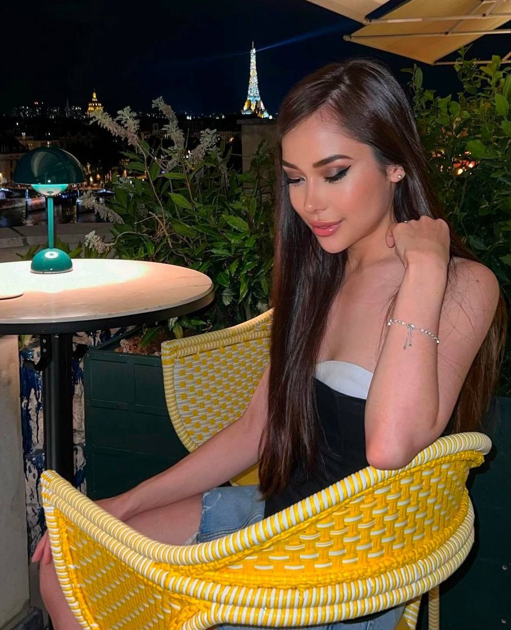 Beautiful brunette fashion model sitting at a luxury Paris rooftop restaurant with the Eiffel Tower illuminated at night, elegant evening outfit, premium travel lifestyle photography