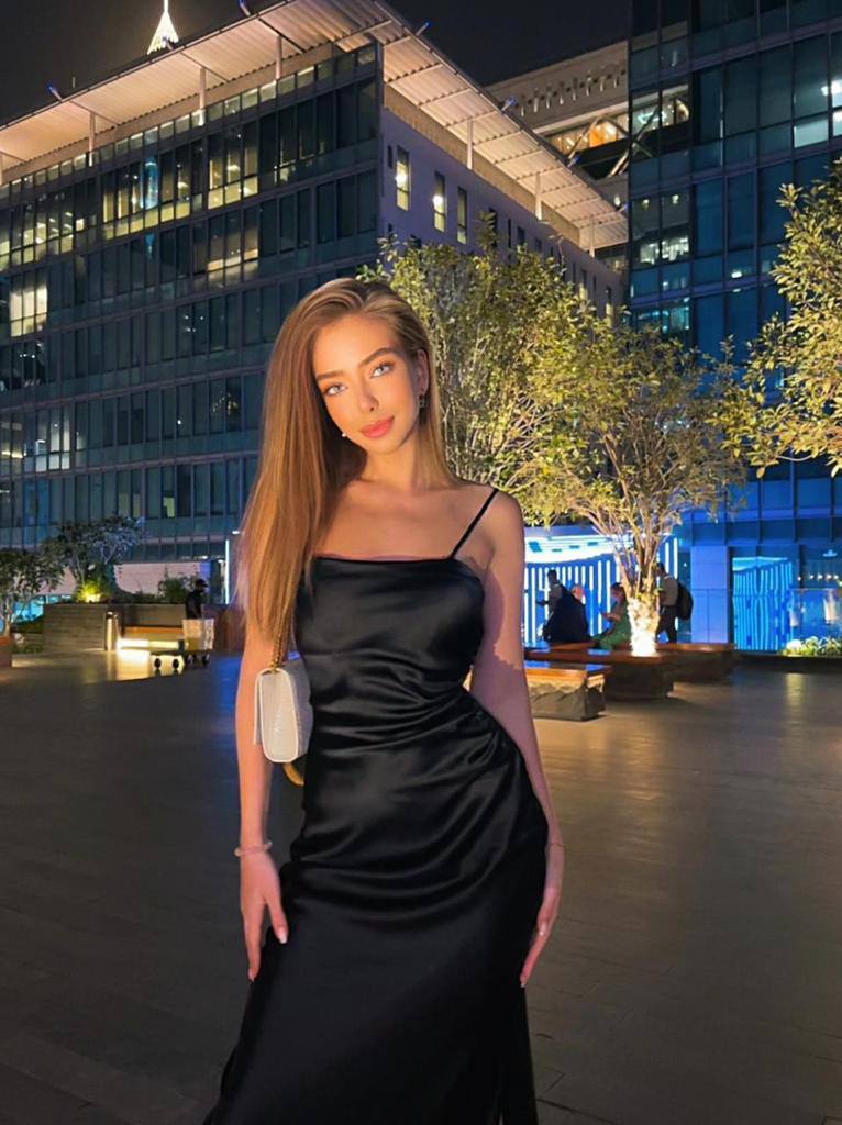 Blonde woman in a black satin slip dress with a small white bag standing on a plaza among illuminated buildings at night