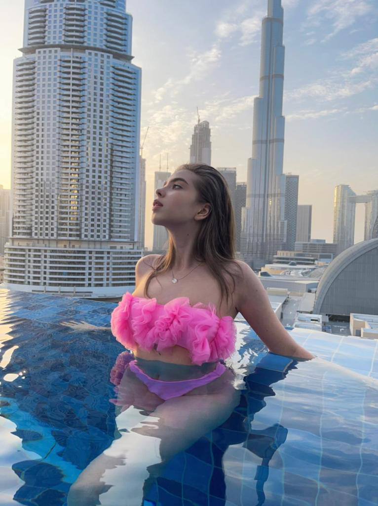 Young woman in a pink ruffled tulle bandeau bikini relaxing in a rooftop infinity pool with the Burj Khalifa and Dubai skyline behind
