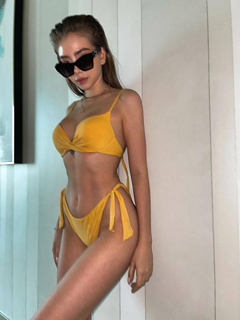 Woman in a yellow tie-side bikini and oversized black sunglasses posing against a white striped wall