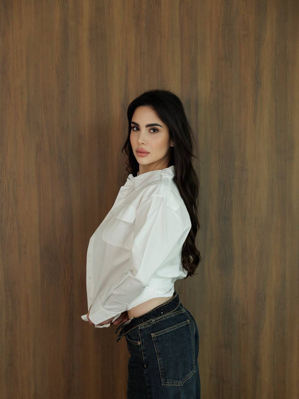 Luxury brunette escort model wearing an oversized white shirt and jeans in a modern lifestyle portrait