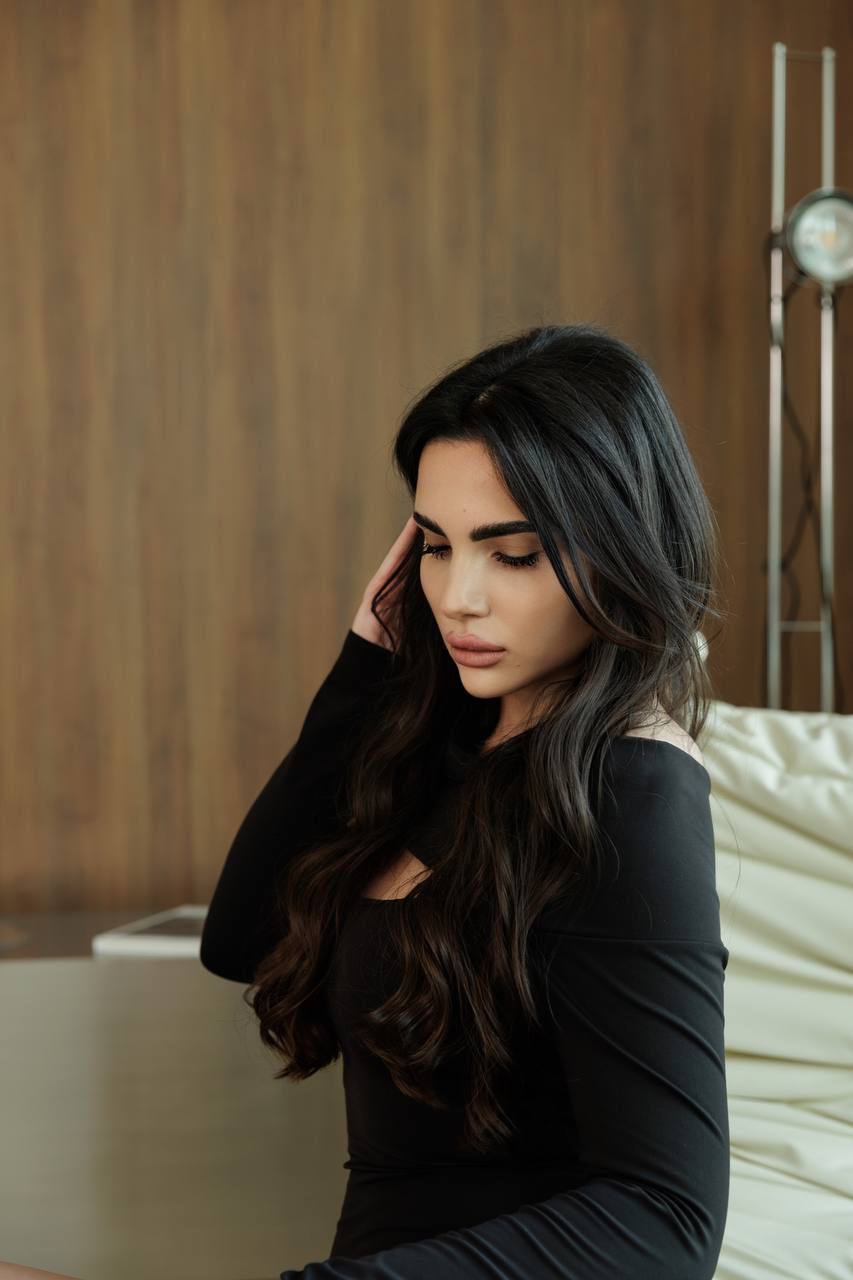 Luxury escort model in a stylish black dress sitting indoors during an elegant lifestyle photoshoot