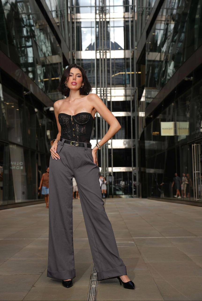 Stylish brunette fashion model with sleek bob haircut wearing a black lace corset and wide-leg gray trousers posing in a modern glass business district with luxury editorial photography