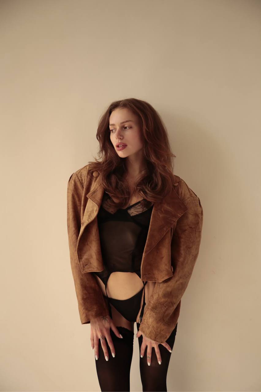 Elegant brunette fashion model wearing sheer black lingerie with a vintage brown leather jacket posing against a minimalist beige background