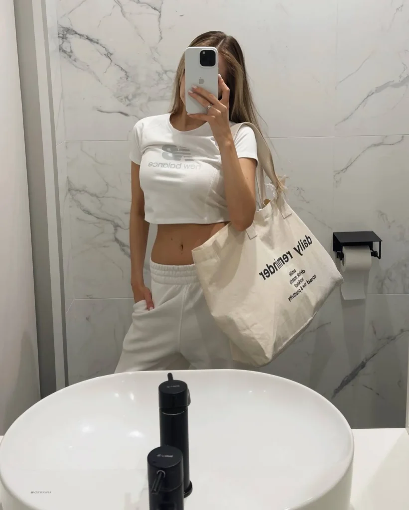 Mirror selfie of a blonde woman in white crop tee and white sweatpants with a canvas tote bag in a marble bathroom