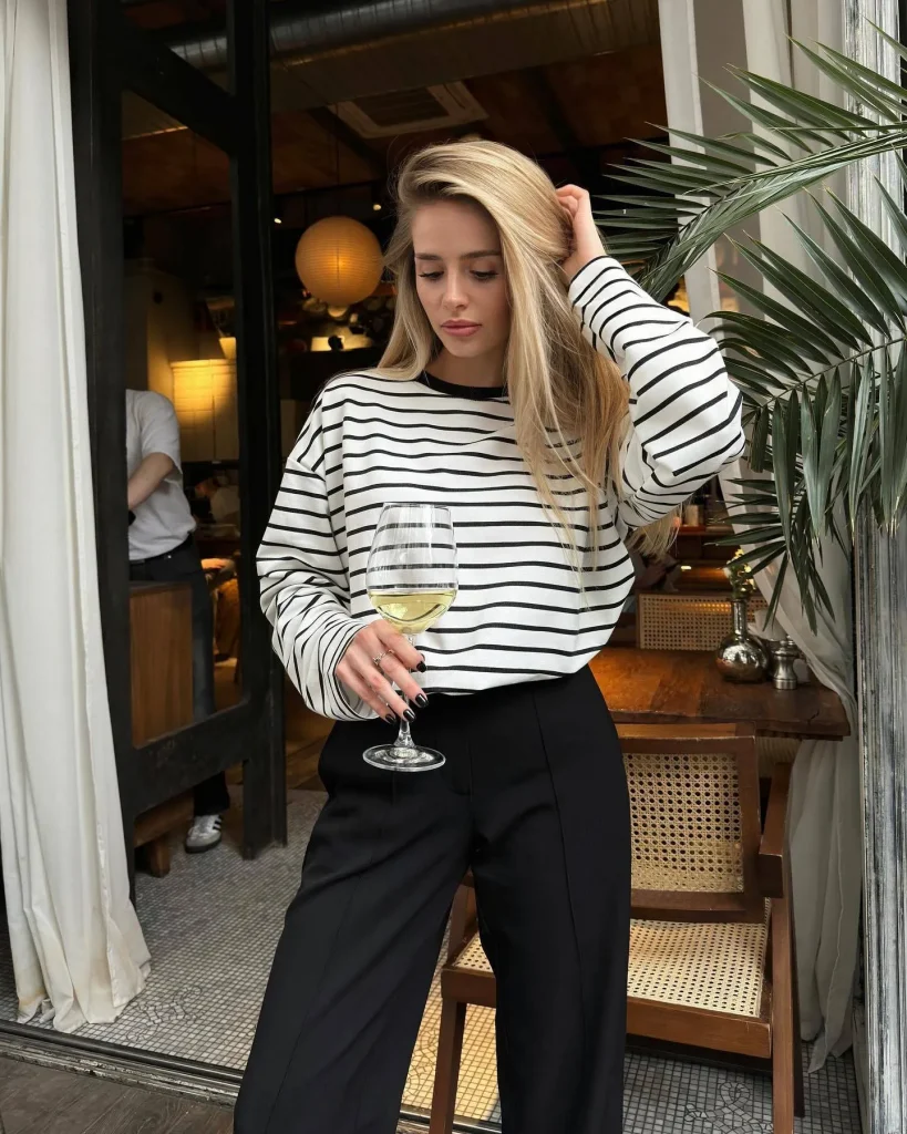 Blonde woman in striped top and black trousers holding a glass of white wine at a cafe entrance