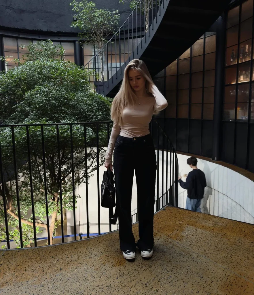 Blonde woman in beige fitted top and black wide-leg jeans with a black bag standing on a curved walkway