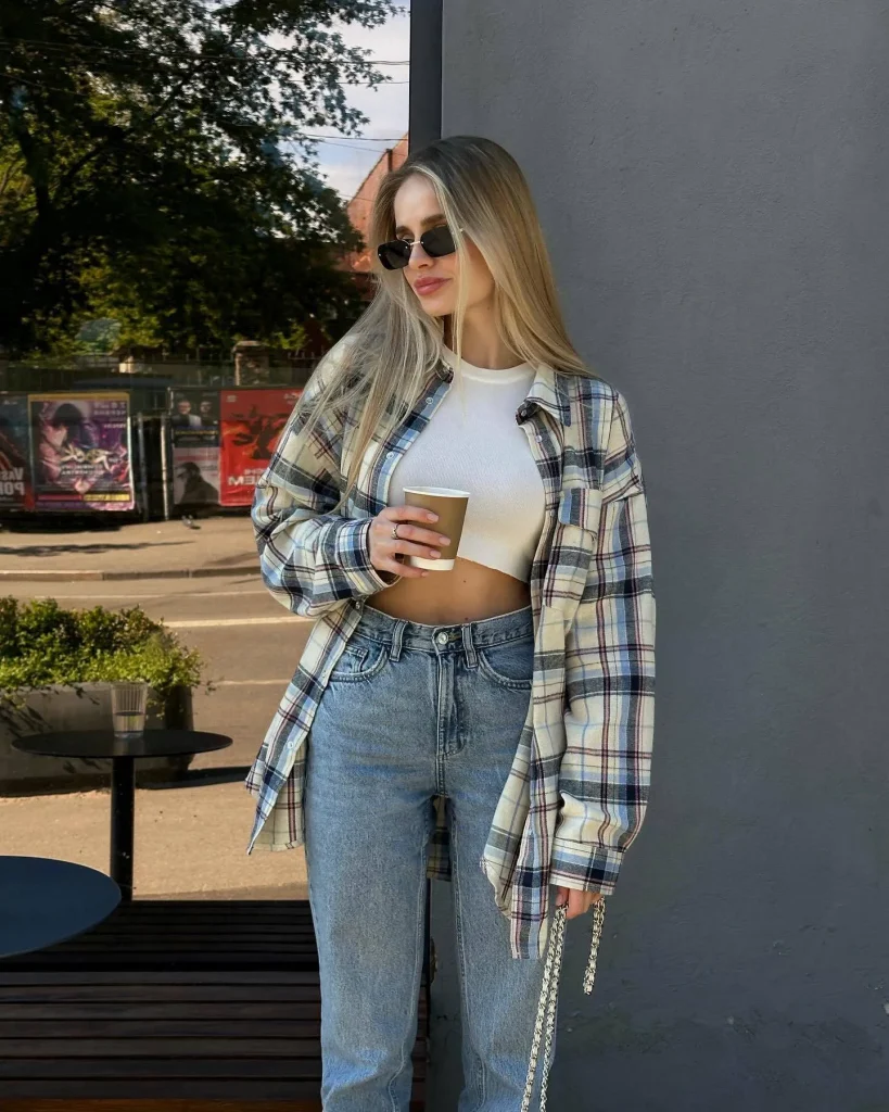 Blonde woman in oversized plaid shirt, white crop top and blue jeans holding a coffee cup on a city street