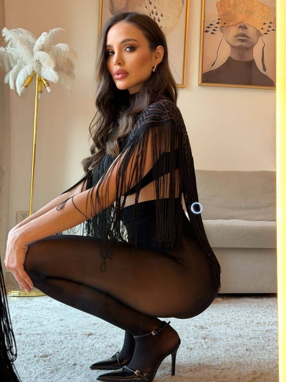 Fashion portrait of brunette woman in a black fringe bodysuit with sheer tights posing in a modern luxury living room
