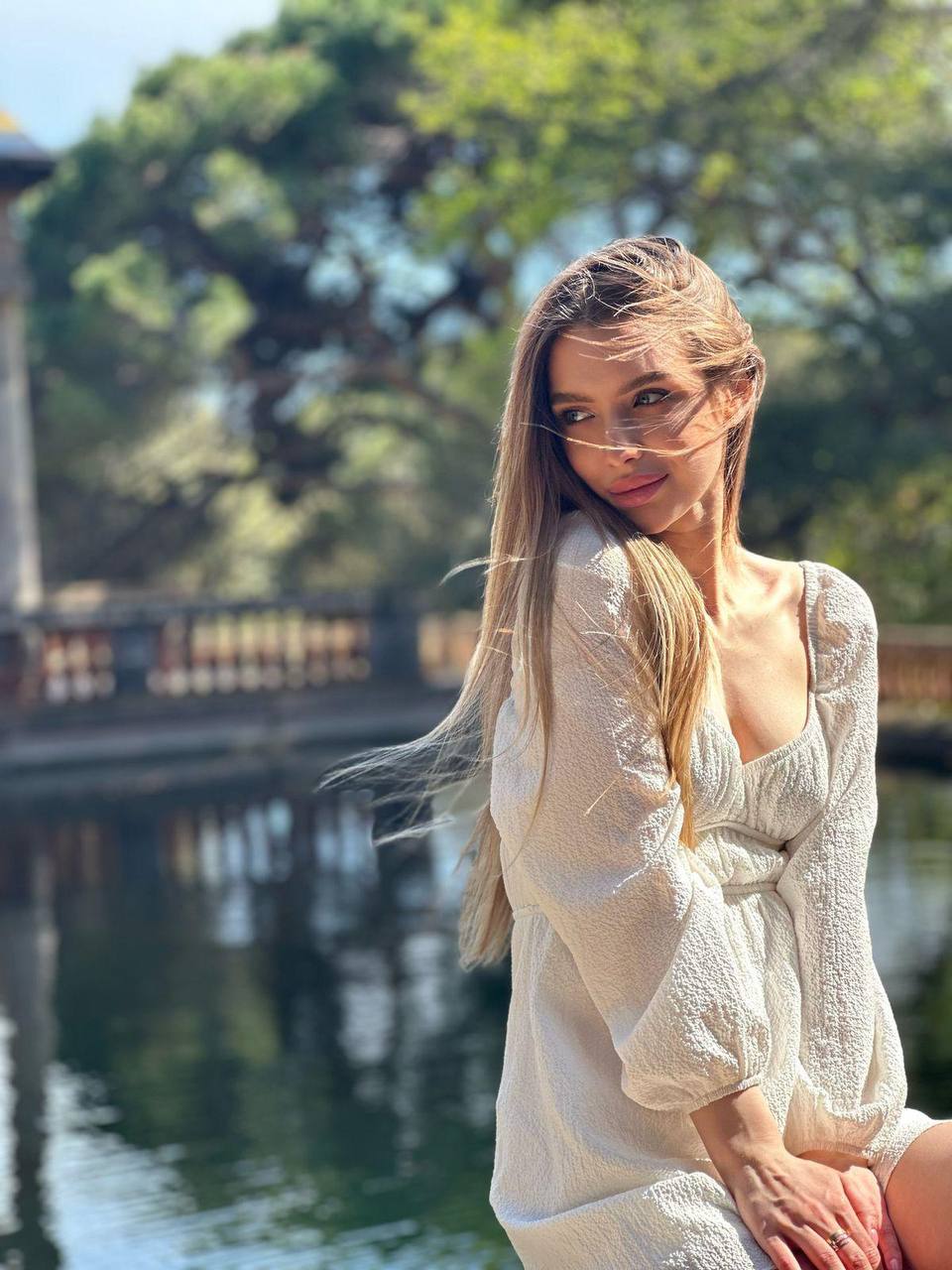 Elegant blonde woman in a white long sleeve dress posing beside a peaceful lake with soft natural sunlight