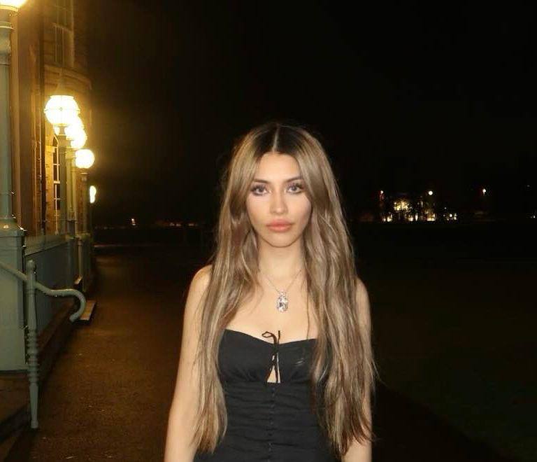 Beautiful blonde fashion model with extra long straight hair wearing a black evening dress posing outdoors at night under vintage city lights with an elegant editorial style