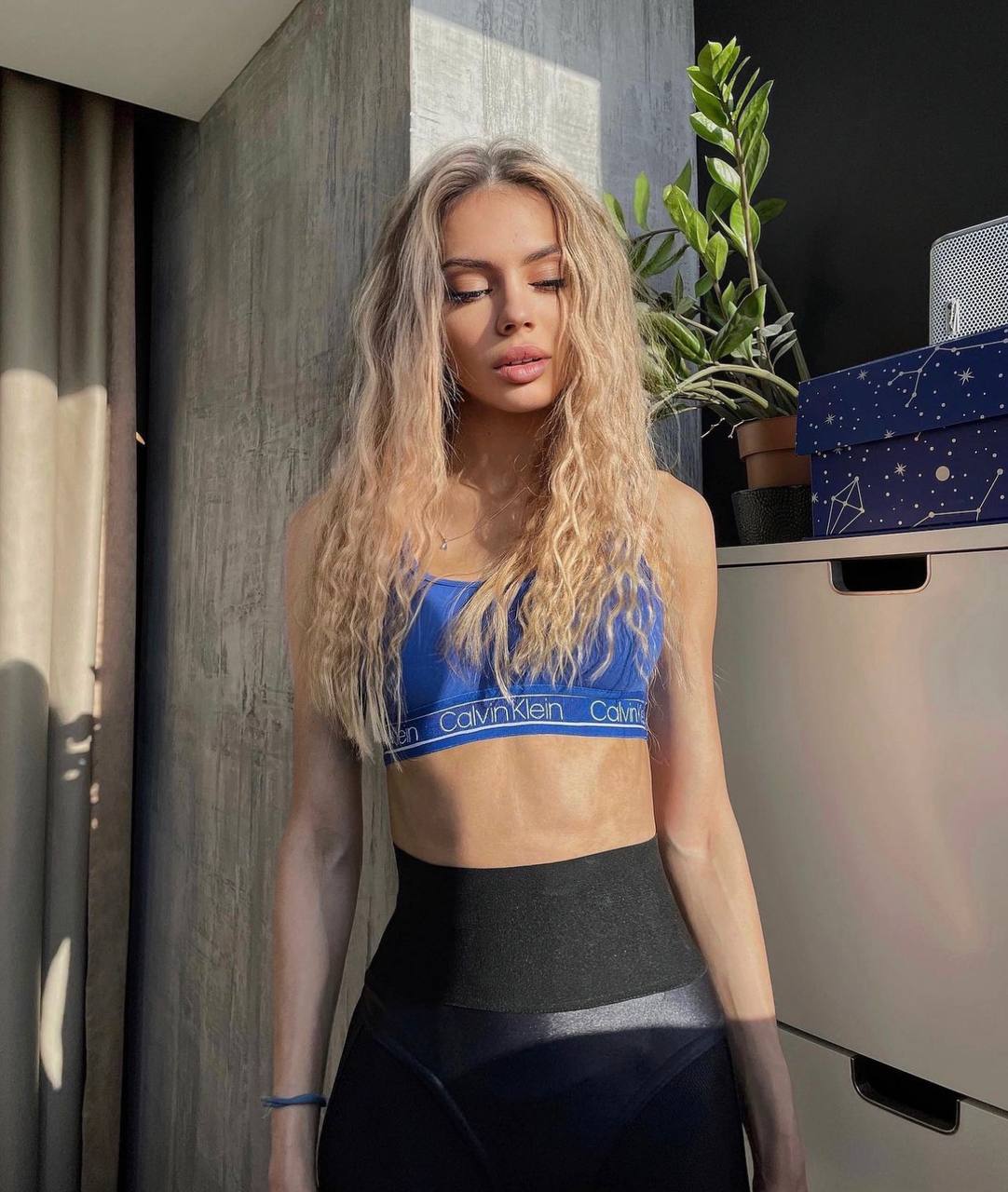Blonde woman with long wavy hair in a blue sports bra and black leggings standing in sunlight next to a green houseplant