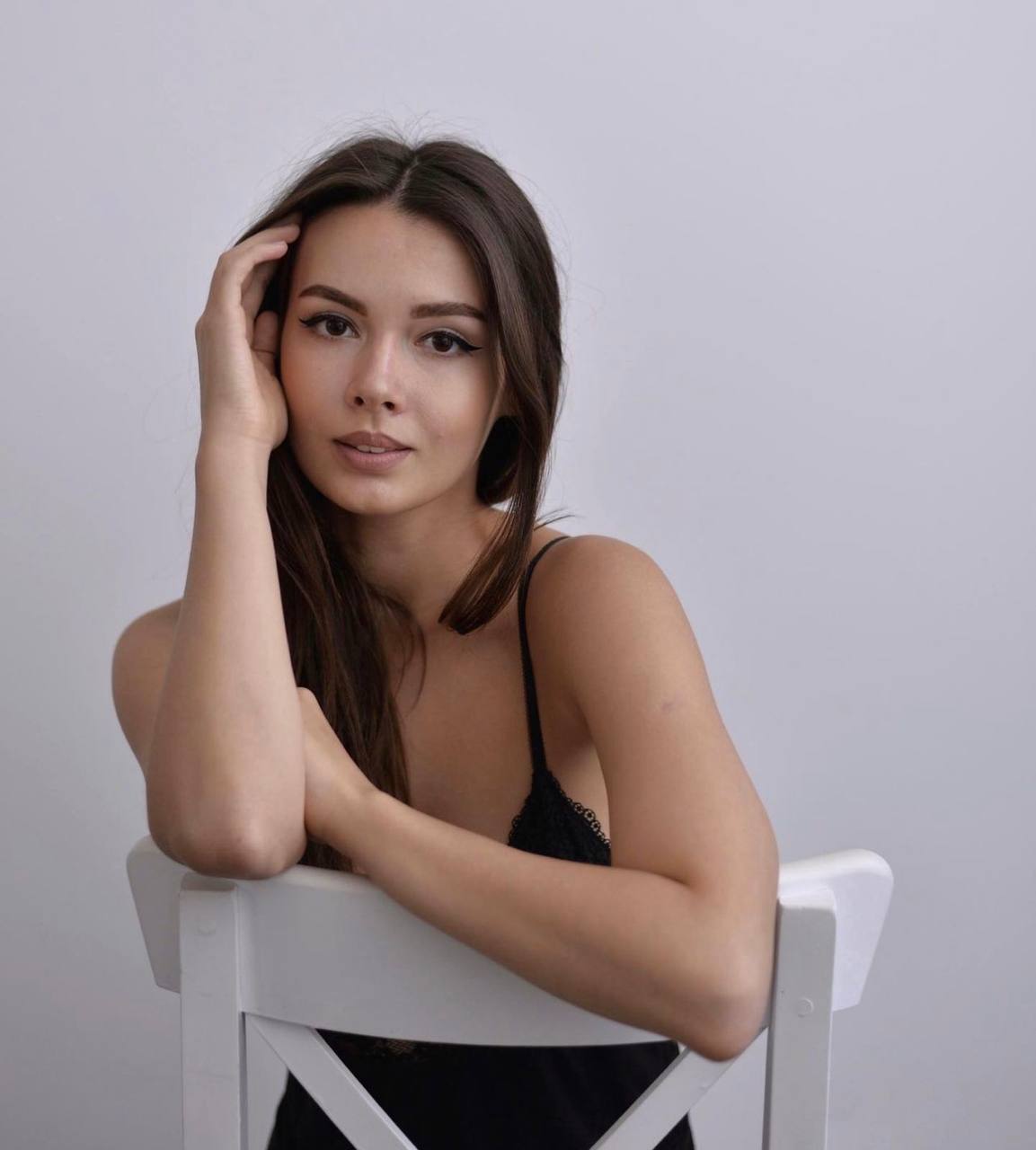 Studio portrait of a brunette woman in a black lace strap top sitting backwards on a white wooden chair and leaning on her arms