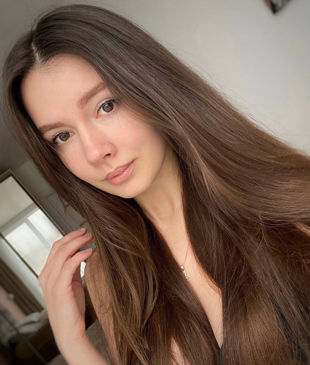 Close-up selfie of a young woman with long straight brown hair, natural makeup and a delicate necklace in soft daylight
