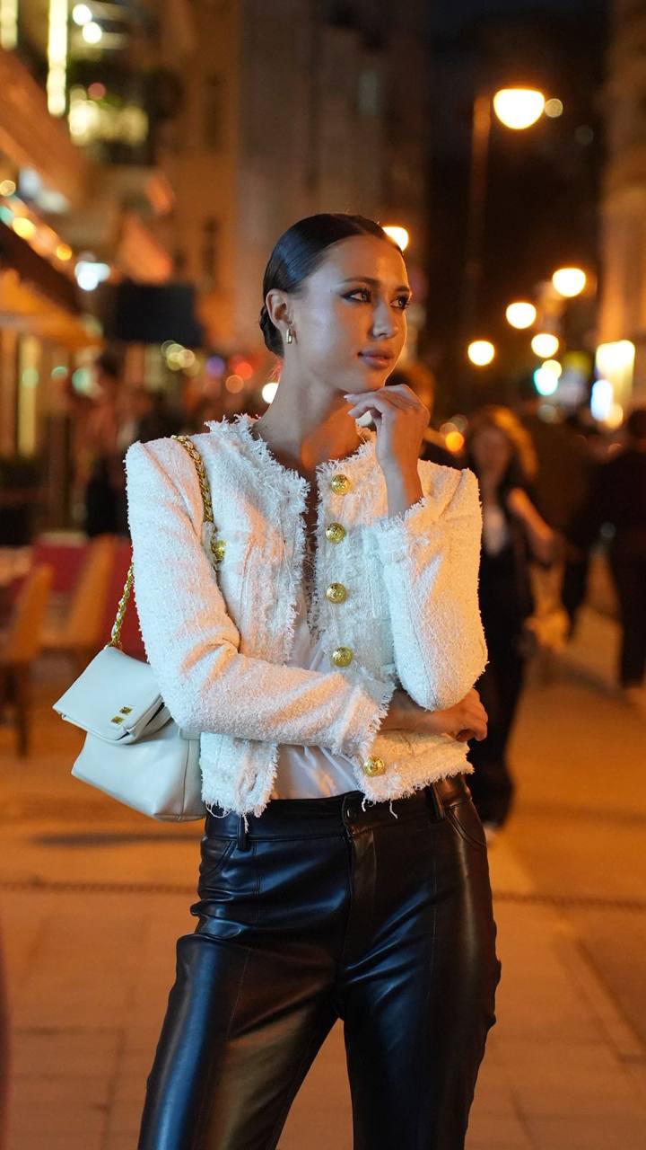 Luxury fashion portrait of a stylish brunette model wearing a white tweed blazer and black leather pants on a city street at night.
