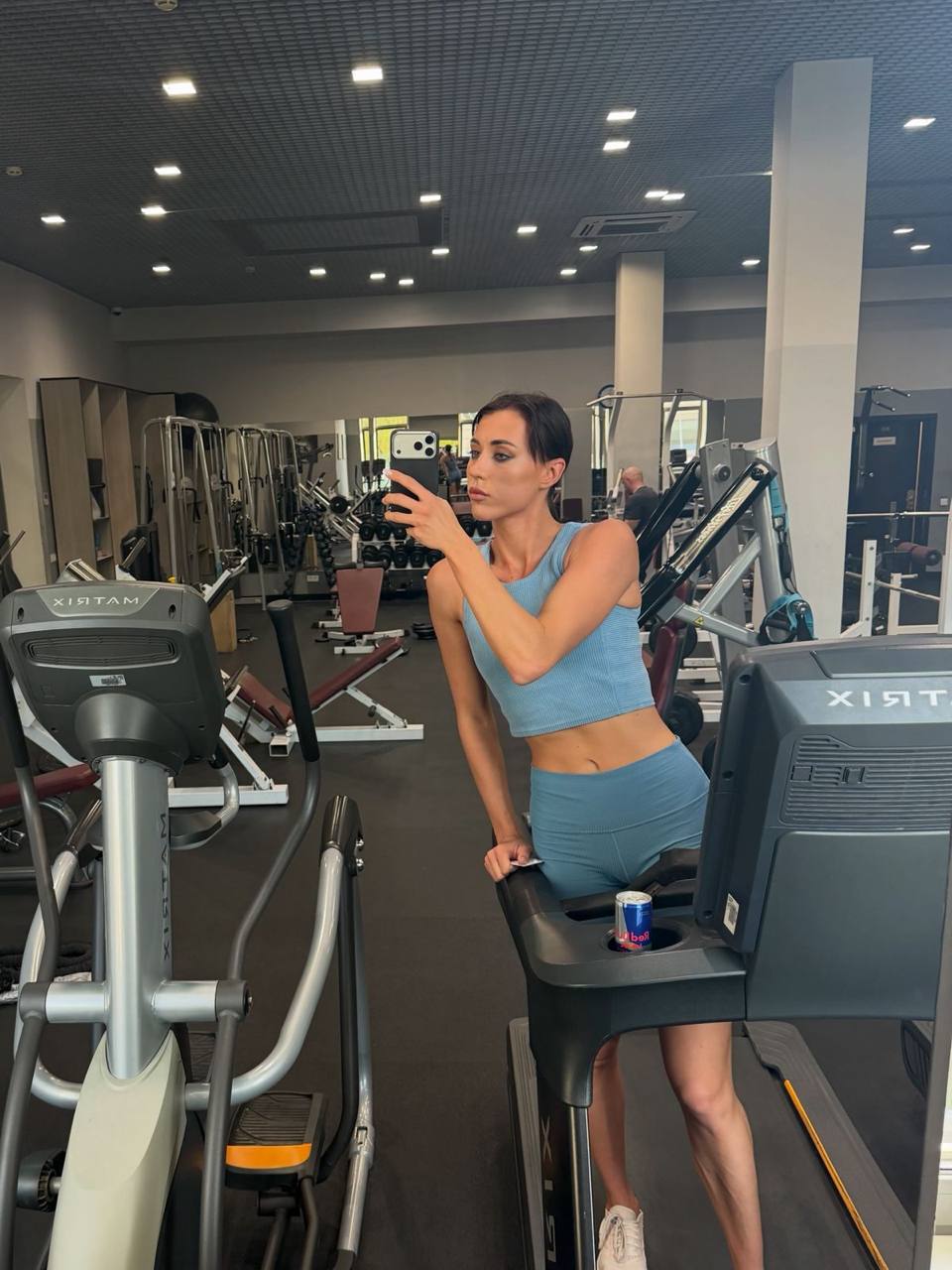 Fitness model posing in a modern gym mirror wearing a light blue seamless workout set beside cardio equipment.