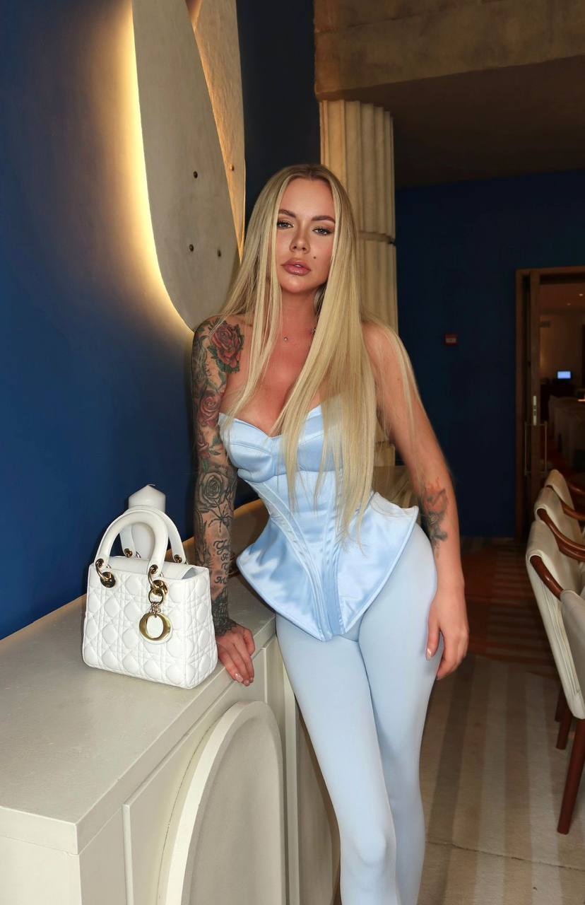 Luxury blonde fashion model wearing a blue corset outfit with a designer handbag in a modern interior.