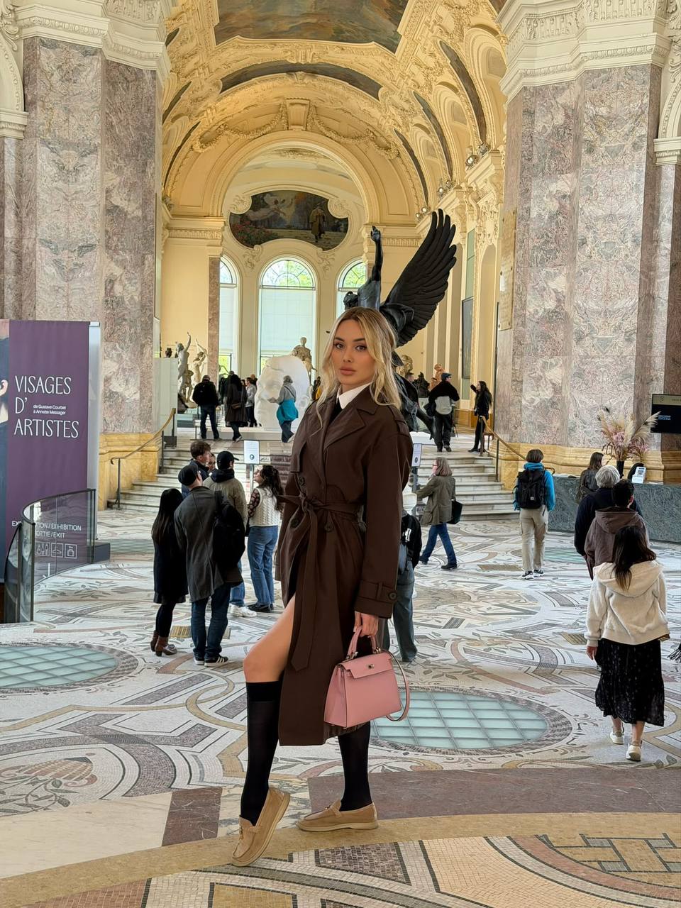 Elegant blonde fashion model wearing a brown trench coat posing inside a grand European museum with classical architecture.