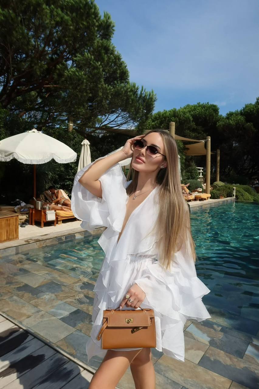 Beautiful blonde fashion model wearing a white resort dress posing beside a luxury swimming pool holding a designer handbag.