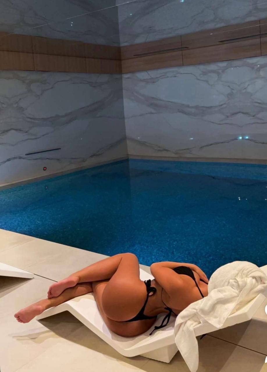 Beautiful woman with long brunette-blonde hair wearing an elegant black bikini, relaxing beside an indoor marble swimming pool inside an ultra luxury spa, soft ambient lighting, expensive wellness resort, calm premium atmosphere, editorial lifestyle photography, ultra realistic.