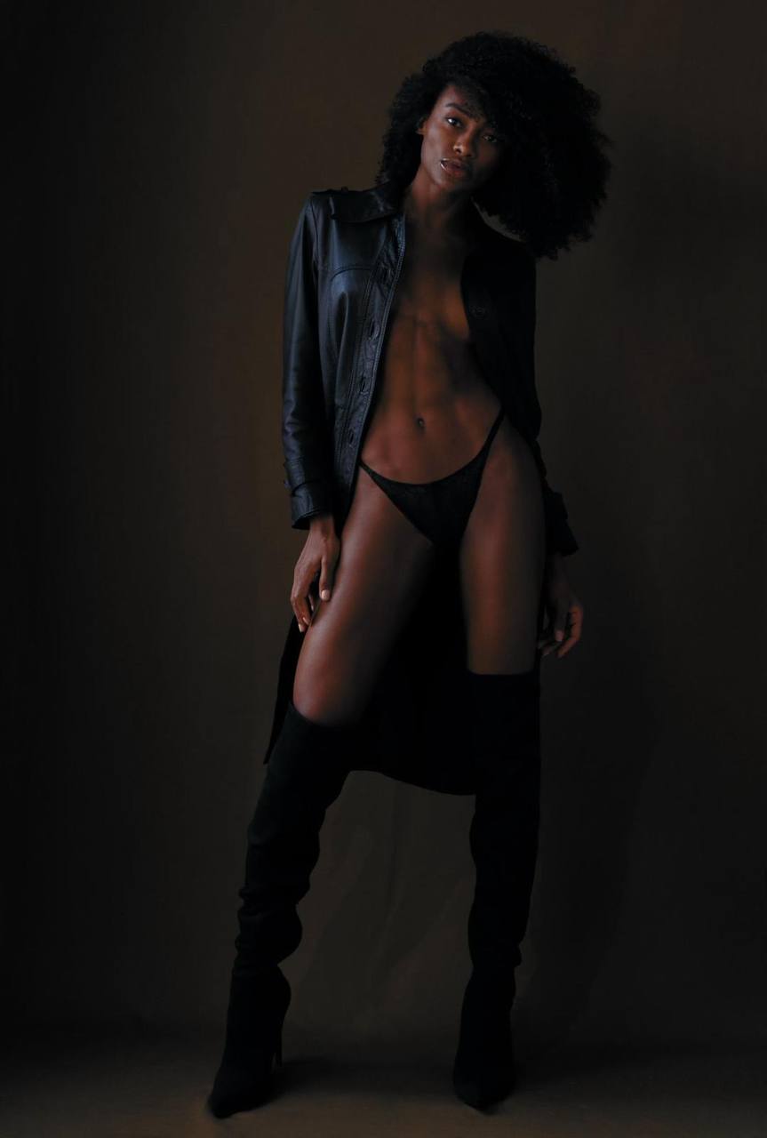 High fashion editorial featuring beautiful Black model wearing black leather coat and thigh high boots in dramatic low light studio